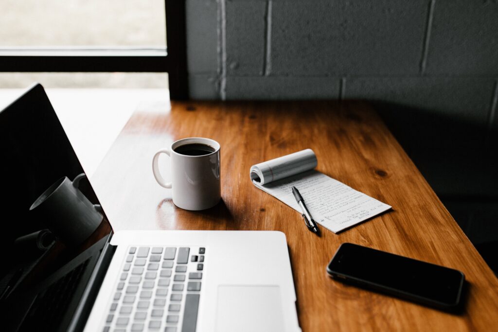 A laptop, cup,notepad and phone on a brown table